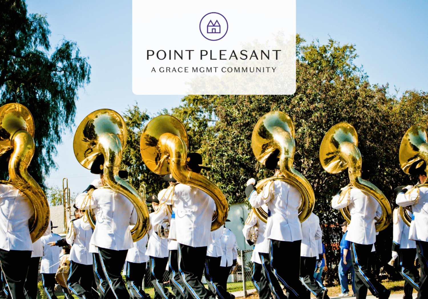 Marching band in white uniforms playing large brass tubas outdoors, with a Point Pleasant logo banner above.”,