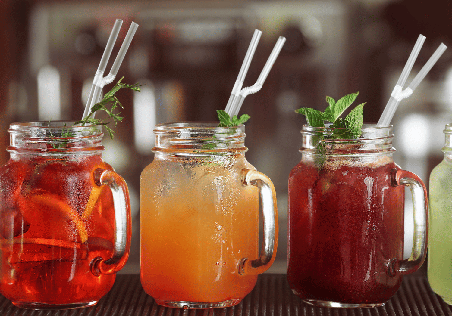 Three mason jar cocktails with mint garnishes and white striped straws on a bar surface.