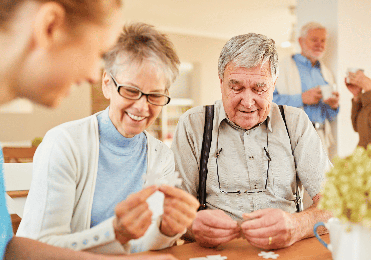 Smiling elderly couple playing cards at a table with a caregiver nearby in a bright, social setting.