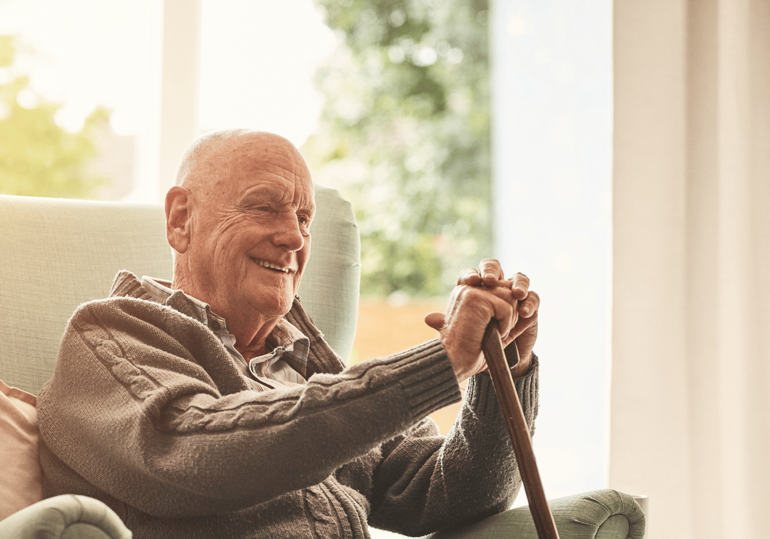 Elderly man smiling while seated in a chair, holding a wooden walking cane with both hands indoors by a sunlit window