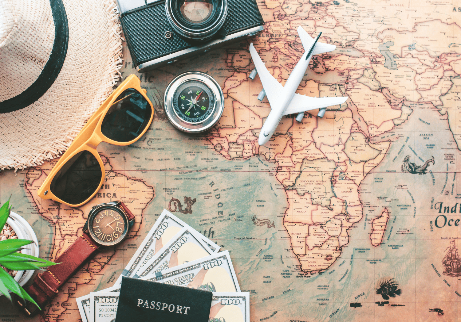 Travel setup on a map with a camera, compass, toy airplane, straw hat, orange sunglasses, passport and cash nearby.
